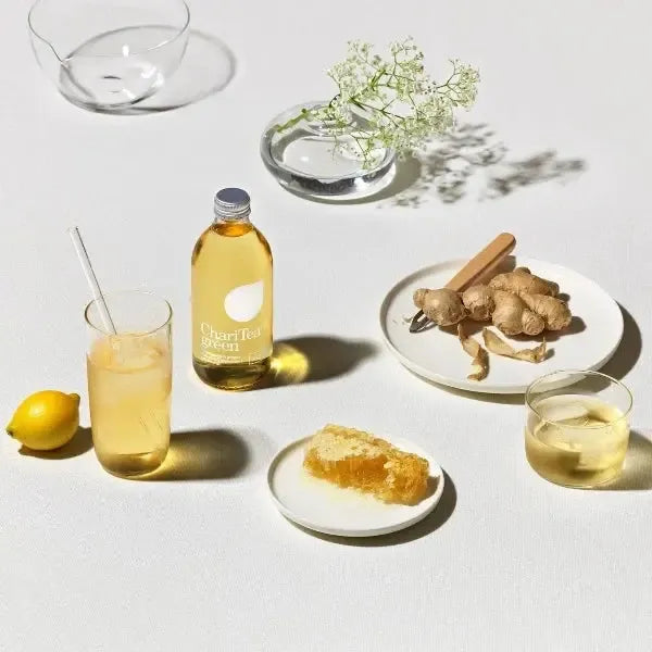Bottle of Charitea Iced Green Tea with glasses, lemon, ginger root, and honey on a white surface.