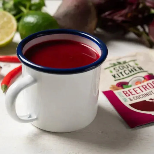White mug containing Soul Kitchen Thai Beetroot Soup, placed on a white surface with vegetables and a 'Soul Kitchen' packet in the background.
