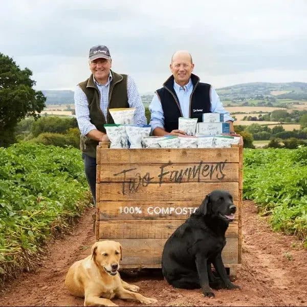 Two farmers with their dogs standing next to a wooden box labeled 'The Farmers 100% Compostable' in a field.