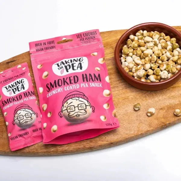 Two pink 'Taking the Pea' snack packages on a wooden board with a bowl of snack mix.